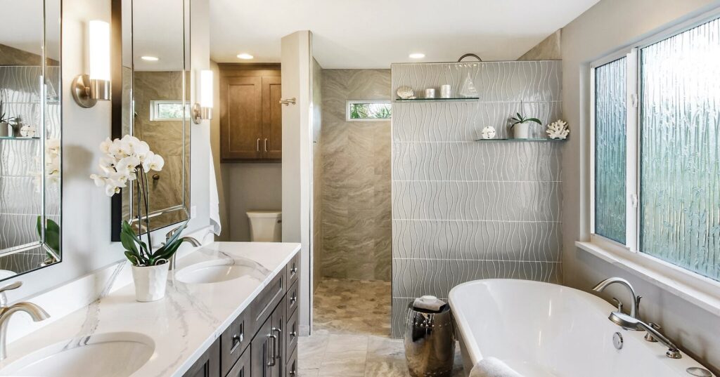 A SandStar Home bathroom project. A 2-sink white marble countertop with dark brown cabinetry sits near a standalone white porcelian bath tub. Frosted glass provides modern appeal.